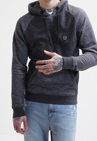 Homme portant un sweat à capuche Volcom gris foncé et un jean bleu clair déchiré, montrant des tatouages sur la main et le cou, sur un fond uni.