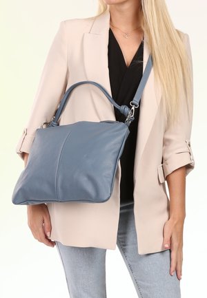 Woman dressed in beige blazer and black top holding a medium-sized blue leather handbag with shoulder strap and top handles.
