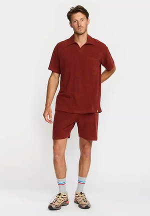 Homme debout portant une chemise à manches courtes et un short assortis bordeaux, des chaussettes grises à rayures et des baskets beiges sur fond blanc.