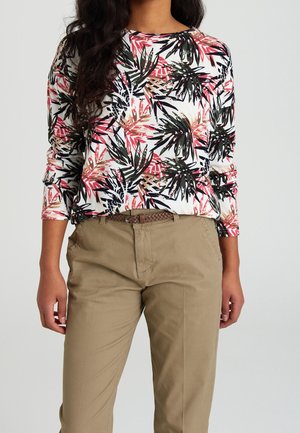 Frau trägt ein weißes Langarmshirt mit schwarzen, pinken und braunen Blattmustern, das in eine beige Hose mit geflochtenem Gürtel gesteckt ist.