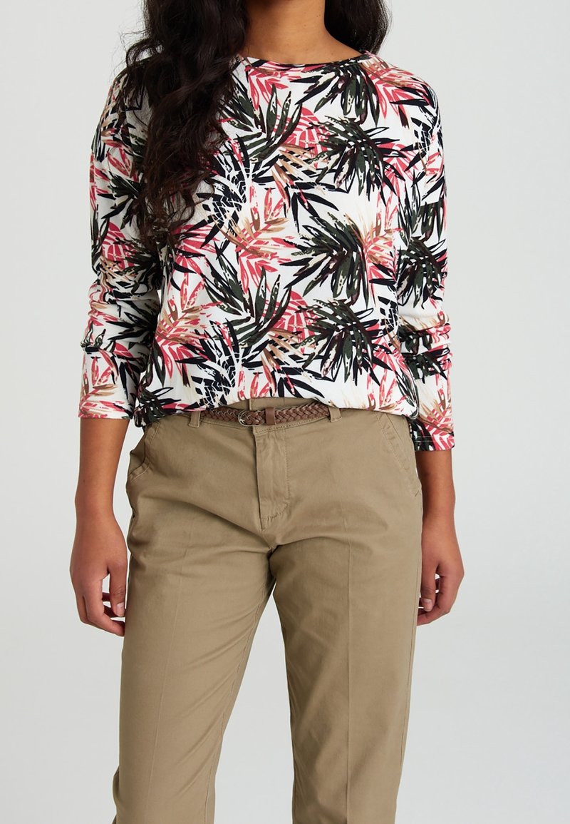 Femme portant un haut blanc à manches longues avec des motifs de feuilles noirs, roses et beiges, rentré dans un pantalon beige avec une ceinture tressée.