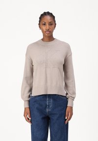 Maglione lavorato a maglia beige con scollo rotondo a coste, vestibilità ampia e logo "CK" in rilievo nella parte anteriore, abbinato a jeans blu scuro.