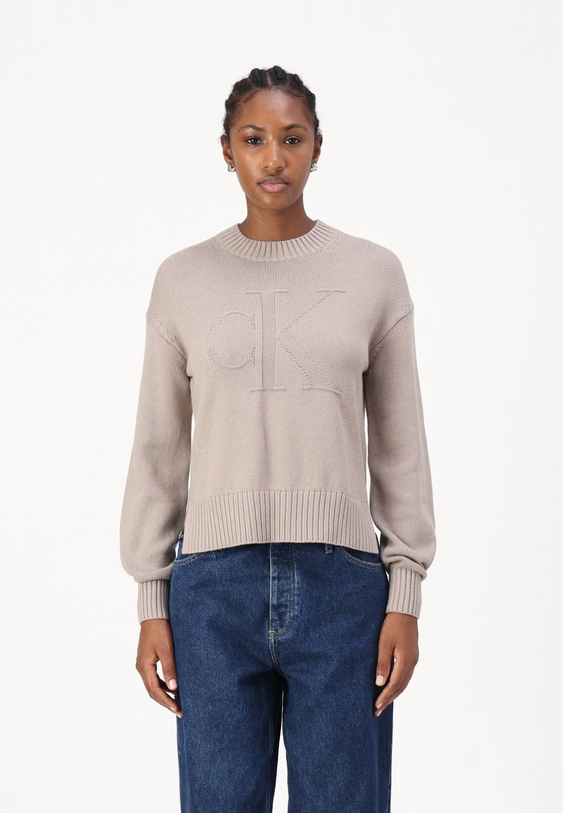 Maglione lavorato a maglia beige con scollo rotondo a coste, vestibilità ampia e logo "CK" in rilievo nella parte anteriore, abbinato a jeans blu scuro.