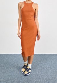 Hladké, bezramenní oranžové šaty s přiléhavým střihem, v kombinaci s modrožlutými teniskami, na osobě na jednobarevném pozadí.