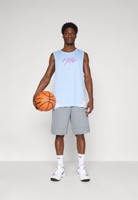 Mann in hellblauem ärmellosem Basketballtrikot und grauen Shorts, der einen orangefarbenen Basketball hält und vor einem einfarbigen Hintergrund steht.