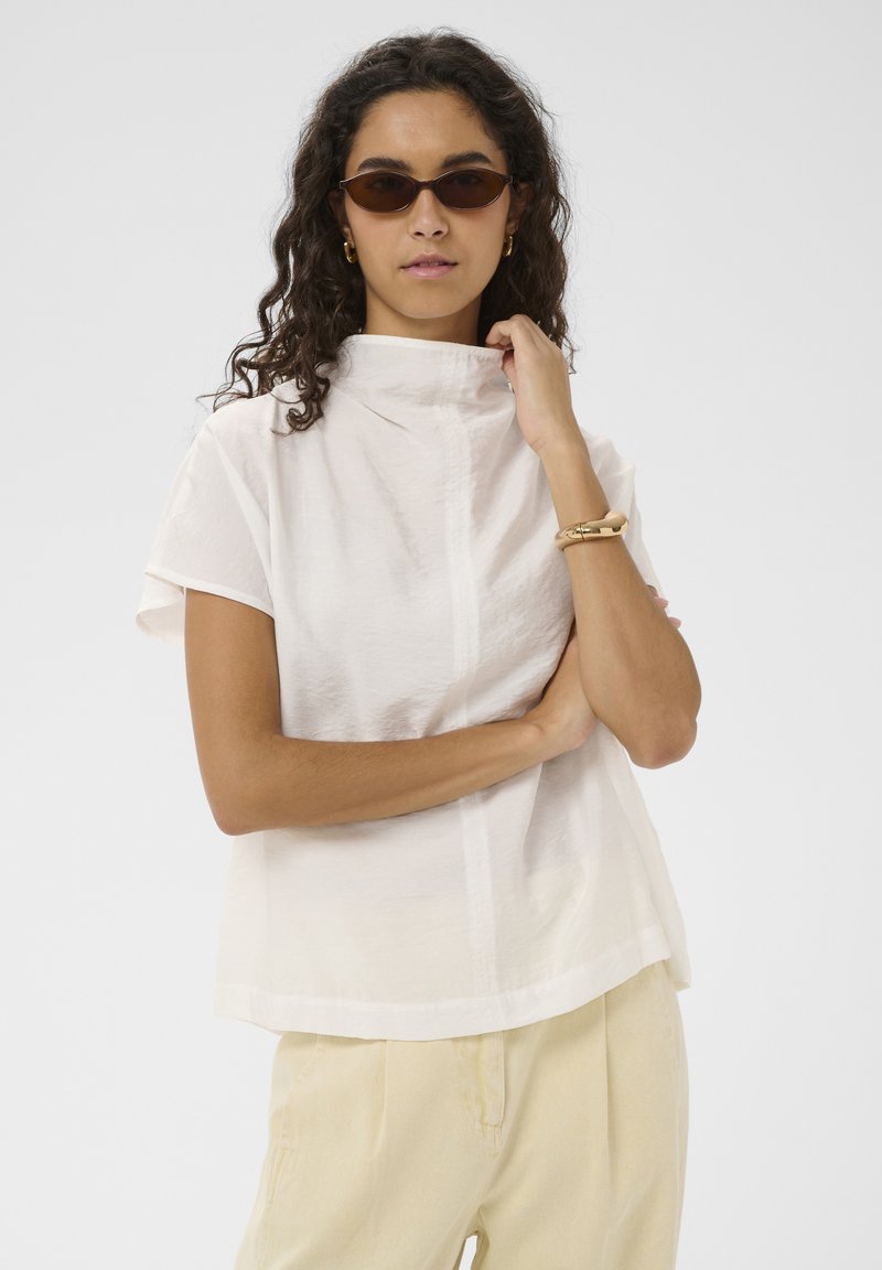 Femme aux cheveux bouclés et foncés, portant des lunettes de soleil marron, un haut blanc à manches courtes avec un col montant, un pantalon beige, un bracelet en or et des boucles d’oreilles créoles.