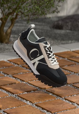 Sneaker neri e beige con lacci bianchi, caratterizzati da un grande logo "CK" sul lato, suola in gomma e materiale superiore testurizzato.