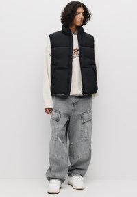 Gilet imbottito nero sopra un maglione bianco con stampa grafica, abbinato a pantaloni cargo grigi chiari. Le sneakers completano l'outfit.