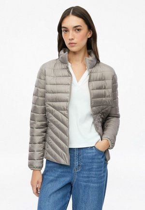 Junge Frau trägt eine hellgraue gesteppte Jacke über einem weißen Hemd und blauen Jeans und steht mit einer Hand in der Tasche.