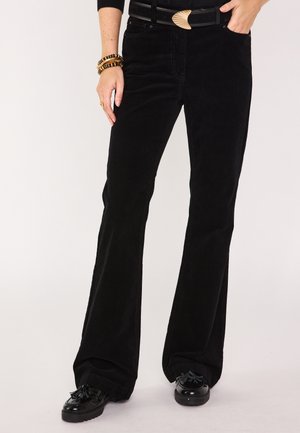 Pantaloni neri in corduroy a gamba larga con tasche frontali e passanti per cintura, abbinati a una cintura nera con una fibbia metallica decorativa. Scarpe nere.