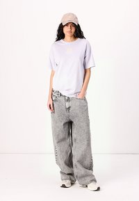 Tricou oversized violet deschis cu mâneci scurte, asociat cu blugi gri largi și o șapcă de baseball bej. Adidașii completează ținuta.