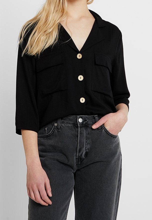 Chemise noire à boutons avec col, deux poches poitrine et trois boutons en bois. Portée avec un jean gris foncé taille haute.