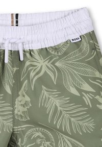 Pantaloni da bagno verdi con vita elastica bianca, chiusura con cordoncino e un design a foglie in verde chiaro. Presentano un’etichetta con logo.