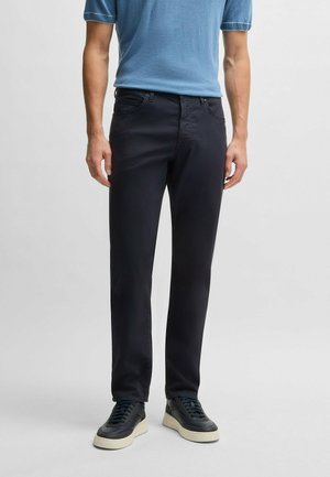 Mężczyzna w czarnych dopasowanych spodniach, czarnych trampkach z białą podeszwą oraz krótkim niebieskim swetrze, stojący na jednolitym tle.