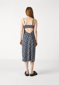 Marineblå blomster midi-kjole med tynne stropper, åpen ryggdetalj og samlet midje. Flate svarte sandaler kompletterer antrekket.