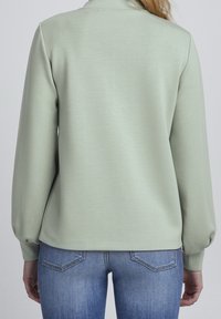 Jersey de color verde claro con mangas largas, textura suave y ajuste relajado, combinado con pantalones vaqueros azules con bolsillos traseros.