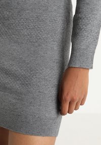 Vestido de punto gris que muestra patrones texturizados y puños acanalados, con un primer plano de la manga y la mano sobre un fondo neutro.