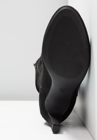 Bottine en suède noir avec bout rond et talon fin. La semelle est en caoutchouc noir texturé pour une meilleure adhérence, avec un design incurvé.