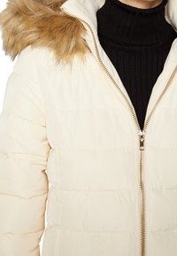 Chaqueta de abrigo en color crema con costuras horizontales. Cuenta con una capucha de piel sintética removible y un cierre de cremallera dorada. Suéter texturizado negro debajo.
