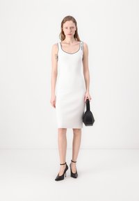 Balts pieguļošs kleita ar apaļu izgriezumu, melni akcenti uz siksnām, apvienota ar melnu ģeometrisku somu un melniem augstpapēžu apaviem.