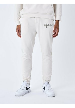 Joggeurs beige clair avec des poignets élastiques et une taille à cordon, arborant un logo gris sur la cuisse gauche. Accouplés avec des baskets blanches.