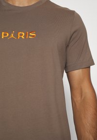 Hnědé bavlněné tričko s krátkým rukávem, s vyšívaným logem "PARIS" a siluetou Jumpman uprostřed.