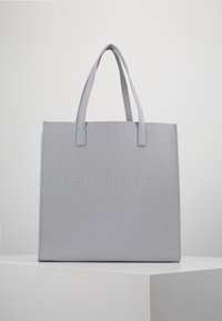 Szara skórzana torba tote z podwójnymi uchwytami, wytłoczona napisem "TED BAKER LONDON" na przodzie, spoczywająca na białym postumencie na jednolitym tle.