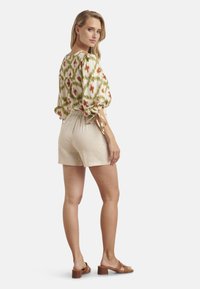 Bloempatronen blouse met pofmouwen, gecombineerd met beige shorts. Model draagt bruine hakken en staat met de rug naar de camera gekeerd.