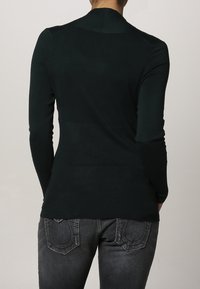 Mørkegrøn ribbet langærmet top med en tætsiddende silhuet og lige halsudskæring, parret med mørke denimjeans med stikninger på baglommerne.