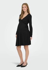 Femme enceinte portant une robe portefeuille noire à manches longues et des chaussures noires à talons bas, debout devant un fond clair uni.