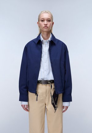 Młoda osoba z blond włosami, ubrana w granatową kurtkę na jasnoniebieską koszulę w paski oraz beżowe spodnie z czarnym paskiem jako dodatkiem.