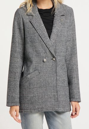 Femme portant un blazer croisé à carreaux gris sur une chemise noire et un jean gris clair, debout devant un fond blanc.