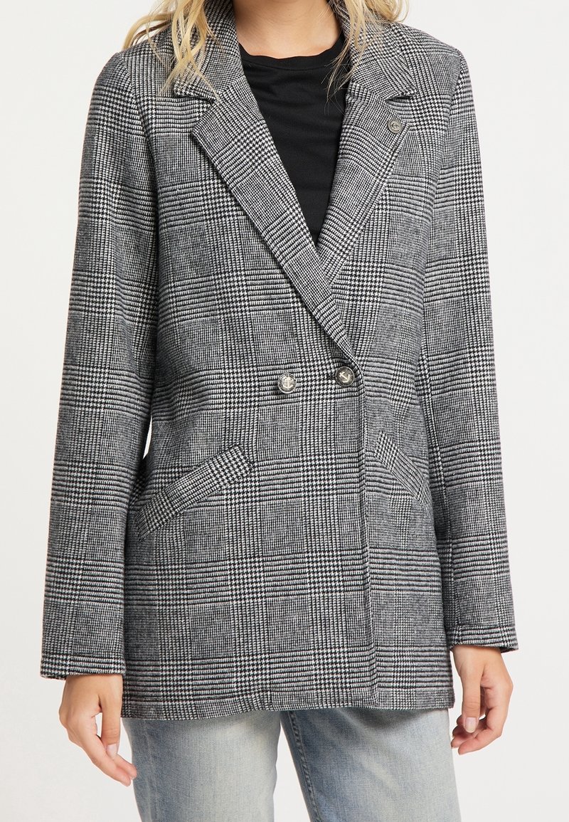 Femme portant un blazer croisé à carreaux gris sur une chemise noire et un jean gris clair, debout devant un fond blanc.