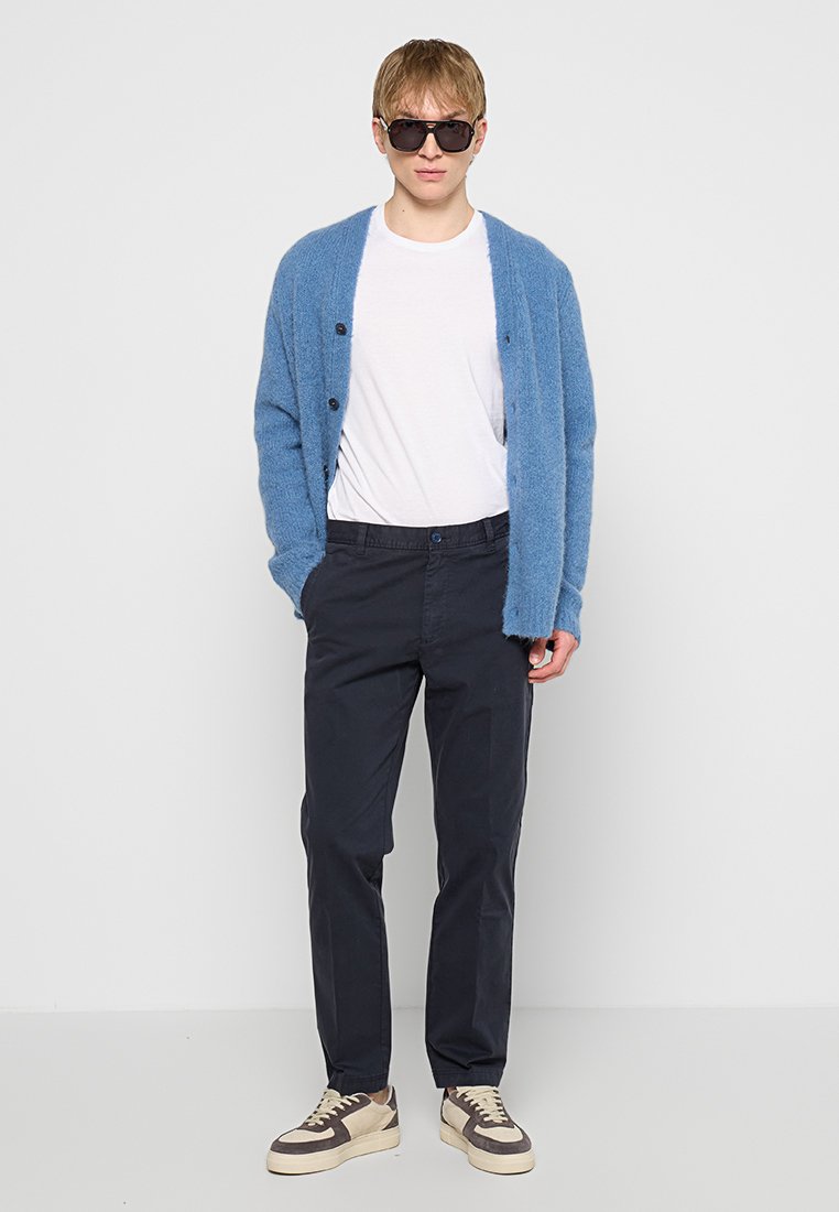 Blauer, flauschiger Cardigan mit Knopfverschluss, weißes T-Shirt, dunkle Hose und beigefarbene Sneakers, ergänzt durch dunkle Sonnenbrille.