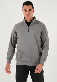 Šedá mikina na zip s vysokým límcem, žebrovanými manžetami a jemným logem na hrudi. Měkká tkanina s hladkou strukturou.