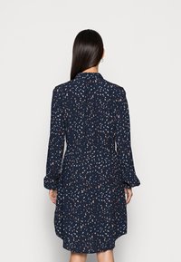 Donna con lunghi capelli scuri che indossa un abito blu navy con chiazzette bianche e beige, in piedi di fronte a uno sfondo bianco semplice.