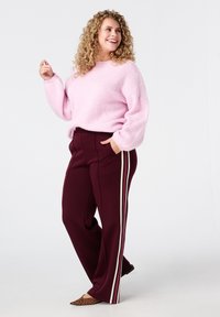 Roze gebreide trui, kastanjebruine wijde broek met witte zijstrepen en leopoardprint hakken. Model heeft krullend haar en staat met een glimlach.