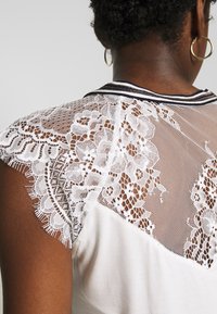 Biały top z koronkowymi i siateczkowymi panelami na ramionach, z okrągłym dekoltem i kontrastowym wykończeniem wzdłuż dekoltu.