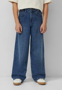 Szerokie niebieskie jeansy z długimi nogawkami mają wysoki stan, pięć kieszeni i subtelne wyblakłe wykończenie. W połączeniu z białymi trampkami dla kontrastu.