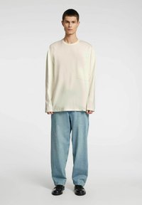 Langermet kremfarget skjorte med brystlomme og lyseblå, vide jeans. Modell står på en nøytral bakgrunn og har på seg svarte sko.