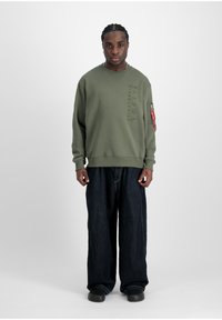 Felpa verde oliva con logo del marchio in rilievo e tasca sulla manica sinistra; abbinata a jeans larghi in denim scuro e scarpe nere.
