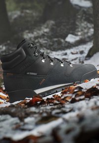 Scarponcino da trekking nero su terreno innevato con foglie cadute, ambiente boschivo all'aperto, vista laterale che mostra i lacci e la suola robusta.
