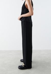 Svart plisserad ärmlös topp i kombination med matchande svarta plisserade vida byxor. Outfiten kompletteras av svarta träskor med gulddetaljer.