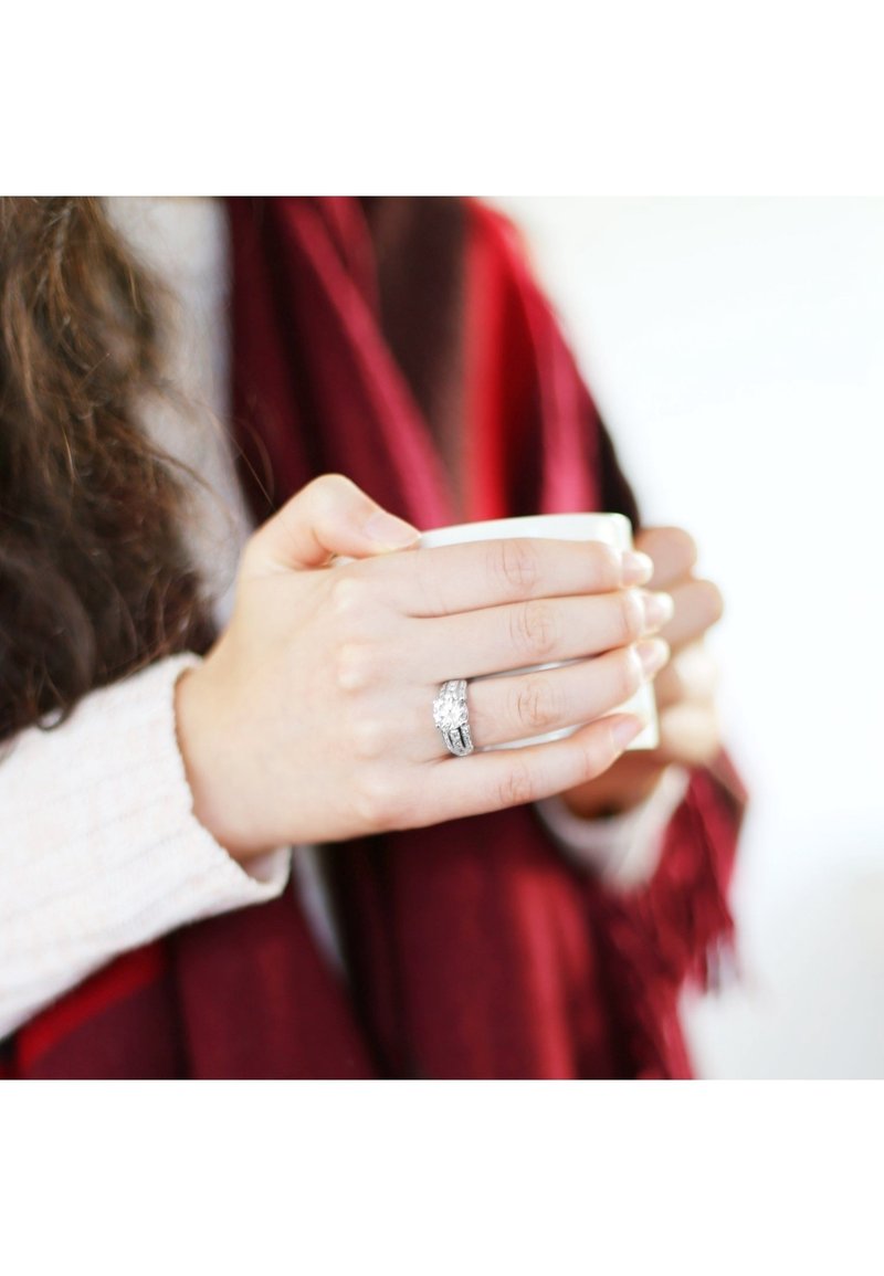Mains tenant une tasse blanche, portant une bague en argent avec une grosse pierre claire et une écharpe rouge visible en arrière-plan.