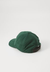 Grønn ullhette med en myk tekstur, buet skygge og et justerbart brunt belte bak. Har innvendige sømmer og ventilasjonsåpninger.
