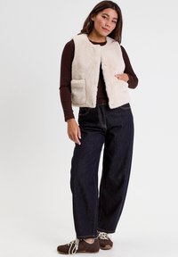Femme portant un gilet en fausse fourrure beige sur un haut marron à manches longues, un jean large foncé et des baskets marron avec des lacets blancs.