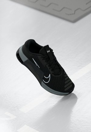 Černá sportovní bota s bílými akcenty, síťovaným svrškem a tlustou polstrovanou podrážkou, která se vznáší nad světlým povrchem s geometrickými vzory.