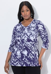 Fioletowa i biała bluzka w wzory z dekoltem w serek i rękawami o długości trzy czwarte, z zapięciem na guziki i luźnym krojem. Odpowiednia do noszenia na co dzień.