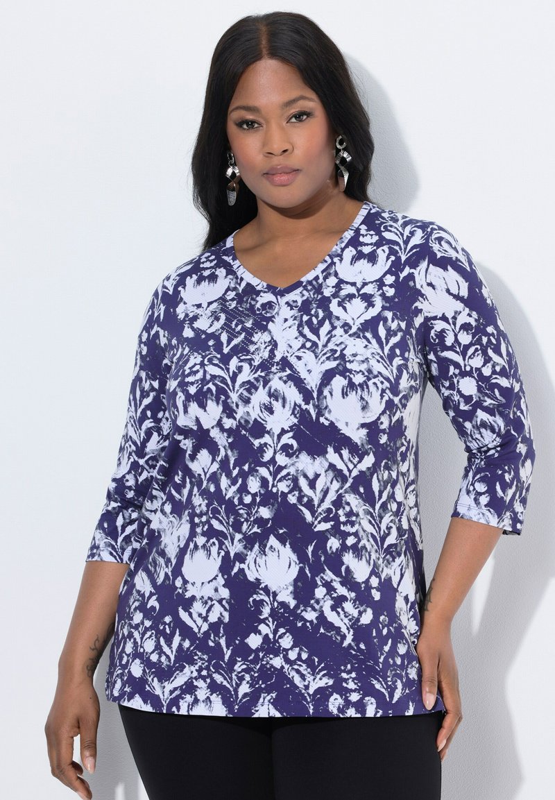 Fioletowa i biała bluzka w wzory z dekoltem w serek i rękawami o długości trzy czwarte, z zapięciem na guziki i luźnym krojem. Odpowiednia do noszenia na co dzień.