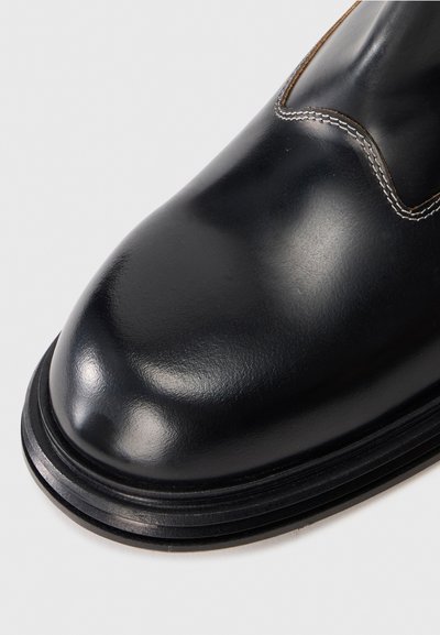 Botte en cuir noir avec un bout arrondi, une texture lisse, des coutures contrastantes et une semelle épaisse noire.